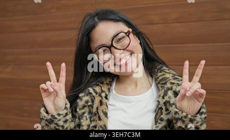Frau blitzt Friedenszeichen mit beiden Händen, trägt Leopardenfellmantel an einer hölzernen Gebäudewand; verspielte Freude. Stockfoto