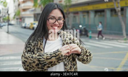 Frau, die mit geschlossenen Augen lächelt, macht Herzform mit Händen auf der Straße, trägt Leopardenmantel und Brille, in der Nähe von Crosswalk und Rollern; Freude Zuneigung. Stockfoto