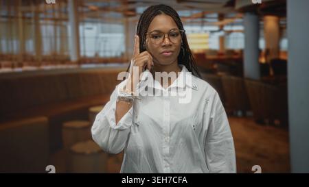 Frau zeigt den Zeigefinger nach oben, während sie in einem dunklen Restaurantgebäude steht und Brillen und Armbänder trägt; Selbstvertrauen. Stockfoto