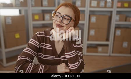 Rothaarige Frau mit Brille auf der Hand neben gestapelten Papppaketen in einem kleinen Verpackungsgebäude; unternehmerische Zufriedenheit. Stockfoto