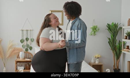 Frau und Mann in Übergröße halten Hände, die neben dem Bett in einem hellen Schlafzimmer tanzen, lächeln und umarmen; Liebesgemeinschaft. Stockfoto