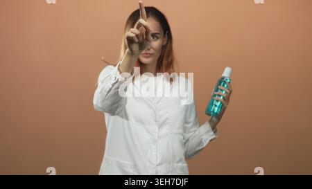 Frau Ärztin und junge Rothaarige im weißen Labormantel hält eine blaue Flasche mit Kappe und zeigt den Zeigefinger in Richtung des Betrachters im Studio; Hygieneanleitung Stockfoto