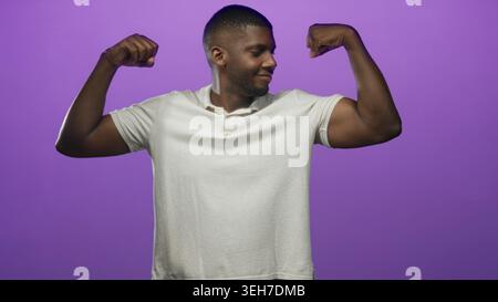 Mann, der im Studio mit violettem Hintergrund Bizeps bicepst, während er lächelnd auf den Arm zeigt; Erfolg der Zuversicht. Stockfoto