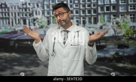 Mann Arzt mit weißem Mantel und Stethoskop zuckt Schultern mit Palmen vor Kanalgebäuden auf einer Straße; Unsicherheit. Stockfoto