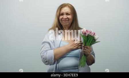 Ältere Frau mit rosafarbenem Tulpenstrauß, Hand auf der Brust und Augen mit Tränenreiz geschlossen, blaues Oberteil und offenes Hemd im Studio; Erinnerung Stockfoto