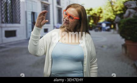 Frau Senior hispanic in hellblauem Tanktop und weißem Cardigan mit orangefarbener Brille hebt die rechte Hand mit gebogenen Fingern und schaut sie auf einem Stree an Stockfoto