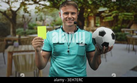 Mann Schiedsrichter hält gelbe Karte und Fußball, trägt Pfeifkopfhörer in Straßencafés; starke Autorität. Stockfoto