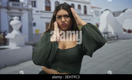 Frau hispanic in Übergröße, die Schläfen berührt und Haare putzt, während sie grüne Bluse und abgeschnittenes Oberteil auf der weißen Stuckstraße trägt; Kopfschmerzen. Stockfoto