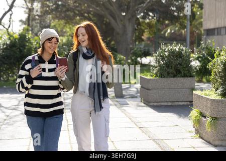 Zwei Studentinnen gehen auf dem Campus, schauen sich das Telefon an, tragen Rucksäcke und Beanie. Freunde, Universität, draußen, sonnig, Bäume, Schatten, Smartphone Stockfoto