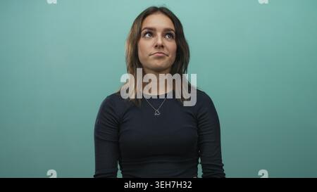 Junge hispanische Frau mit Herzkette blickt im Studio vor blauem Hintergrund auf; nachdenkliche Reflexion. Stockfoto