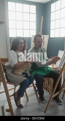 Die Frau zeigt das Smartphone, während der Mann die Farbpalette und den Pinsel im Studio hält, mit einem kleinen Hund auf dem Schoß, der auf die Leinwand zeigt; Konzentration. Stockfoto