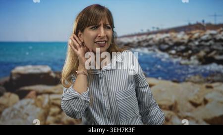 Frau, die das Ohr mit der Hand schröpft und sich nach vorne beugt, mit gestreiftem Hemd und Creolen im Studio Seaside Rocks; Verwirrung beim Hören. Stockfoto