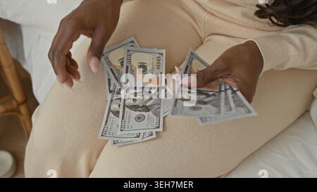 Frau zählt Dollar auf dem Bett, fächert die Hände und sortiert Banknoten im Studio, trägt beige Loungewear und sitzt auf weißen Laken; ruhige Profis Stockfoto