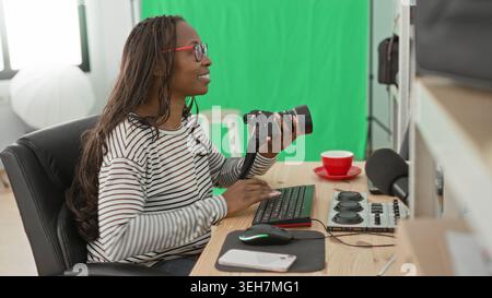 Frau hält Kamera und Typen auf der Tastatur in einem Studio mit grünem Bildschirm und roter Tasse auf dem Schreibtisch; fokussierte Kreativität. Stockfoto