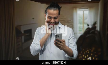 Mann mit Smartphone und Hand an Wange im Wohnzimmer, mit breitem Lächeln und hochgezogenen Augenbrauen auf den Bildschirm; Überraschung. Stockfoto