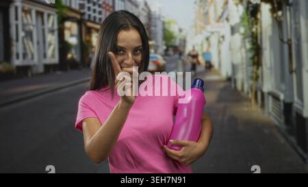 hispanische junge Frau, die eine rosa Flasche in einem Arm hält und mit dem Zeigefinger auf eine Stadtstraße zeigt, während sie lächelt; verspielte Beförderung. Stockfoto