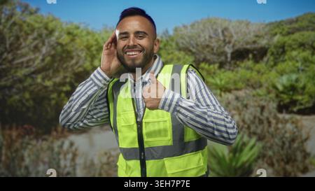 Mann junger hispanic in Neon-Sicherheitsweste umschließt Hand an Ohr und gibt im Wald die Daumen hoch; optimistisch Sicherheit. Stockfoto