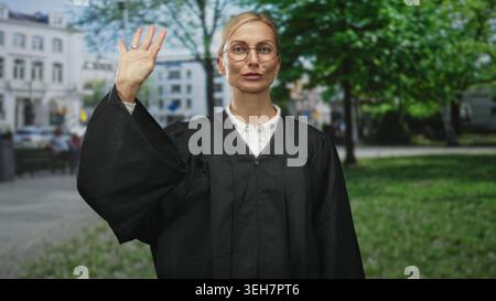 Die Richterin erhebt die rechte Hand mit einer Palme, die vor den Gebäuden der Stadt auf der Straße sichtbar ist, trägt einen schwarzen Justizmantel und eine Brille, während sie leicht lächelt; Autho Stockfoto