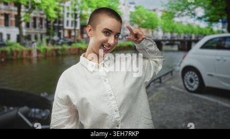 Frau mit kurzen Haaren lächelt und macht ein Friedenszeichen neben ihrem Gesicht neben einem geparkten Auto auf einer Kanalstraße; spielerische Freude. Stockfoto