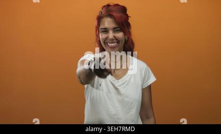 Frau zeigt mit dem Finger auf die Kamera im orangen Studio mit breitem Lächeln und ausgestreckter Armgeste; Freudenverbindung. Stockfoto