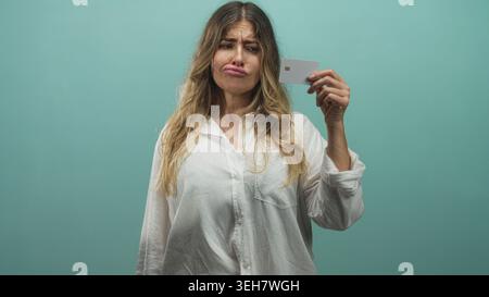 Die Frau zeigt mit dem Finger auf die Kreditkarte, während sie die Karte an der Wand des blaugrünen Studios hält; Frustration Misstrauen. Stockfoto