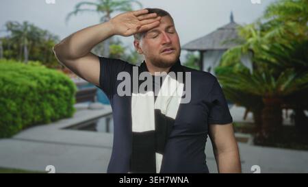 Mann, der die Hand an die Stirn am Pool des Resorts hält, gestreiftes schwarz-weißes Tuch und dunkles T-Shirt trägt, Facepalm-Geste zeigt Kopfschmerzen an; Urlaub fett Stockfoto