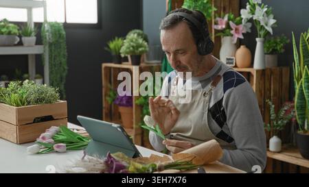 Mann mit Kopfhörern lächelt, Hand auf Tablet, das Tulpen im Blumenladen des Studios arrangiert; Bastelzufriedenheit. Stockfoto