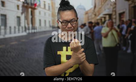 Mann in schwarzem Bitcoin-T-Shirt, der sein Handgelenk umklammert und zuwinkt, während er auf einer belebten Kopfsteinpflasterstraße mit verschwommenen Touristen steht; Unbehagen Krypto curi Stockfoto