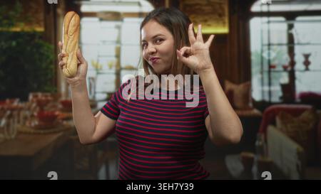 Junge Frau, die ein Baguette mit Handfläche hochhält und Geste im Speisesaal des Hotels in der Nähe von Tisch und Stühlen präsentiert; verspielter Moment. Stockfoto
