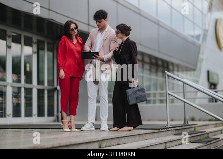 Drei Experten vor einem Bürogebäude, die ein Tablet während eines informellen Geschäftstreffens überprüfen Stockfoto