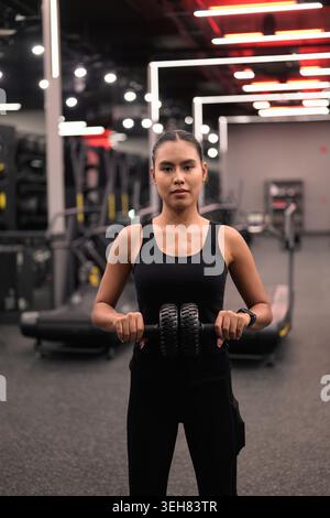 Eine fokussierte junge asiatische Thai-Isan-Frau steht mit einem ab-Rollrad-Übungsgerät und beiden Händen vor der Taille und bereitet sich auf ein Core-Workout vor. Stockfoto