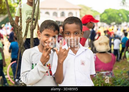 Sri Lanka - 12. Februar 2026: Blick auf zwei lächelnde Schüler, die während einer Veranstaltung im Freien Handgesten machen, mit einer Menge und Grün im Hintergrund. Stockfoto