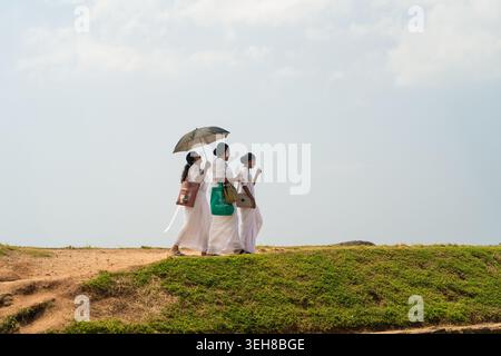 Sri Lanka - 4. Februar 2026: Blick auf drei Frauen in traditioneller weißer Kleidung, die auf einem grasbewachsenen Kamm unter hellem Himmel spazieren, mit einer Frau Stockfoto
