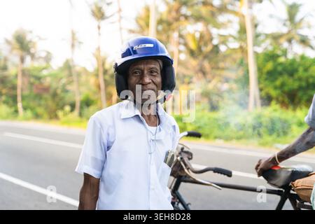 Sri Lanka - 21. Januar 2026: Blick auf einen älteren Mann mit blauem Motorradhelm und weißem Hemd, der auf einer mit Palmen gesäumten Straße neben einem Fahrrad steht Stockfoto