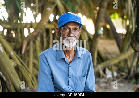 Sri Lanka - 15. Januar 2026: Blick auf einen älteren Mann mit weißem Bart, der eine blaue Mütze und ein Hemd trägt, der vor tropischen Baumwurzeln sitzt. Stockfoto
