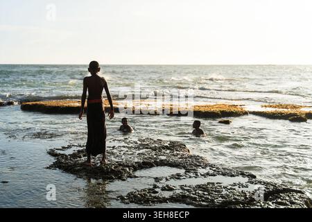 Sri Lanka - 11. Januar 2026: Blick auf Kinder, die während der goldenen Stunde in den flachen Gewässern des Ozeans in der Nähe eines felsigen Riffs spielen. Stockfoto