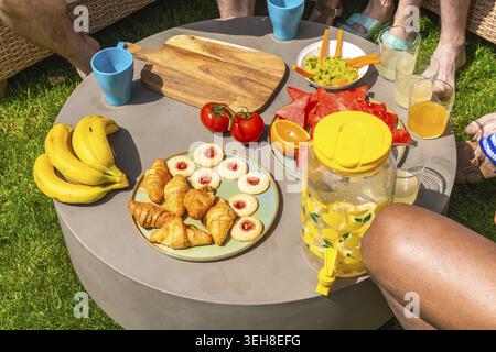 Eine Gruppe von Freunden versammelte sich zu einem zwanglosen Sommer-Picknick und teilte sich an einem sonnigen Tag einen Tisch mit frischem Obst, Gebäck und erfrischendem Zitronenwasser Stockfoto