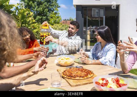 Fröhliche Gruppe von Freunden, die ein sonniges Mittagessen im Hinterhof genießen, Pizza und kalte Getränke teilen, gemeinsam lachen und sich in einer ungezwungenen Outdoor-Umgebung entspannen Stockfoto