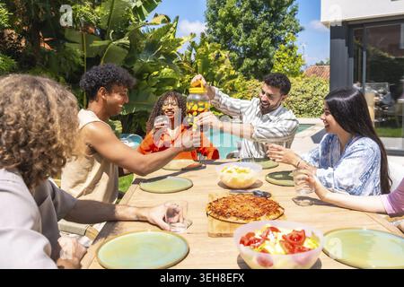 Verschiedene Gruppen junger Freunde lachen und toasten während eines Sommeressens im Freien, mit Getränken, Pizza und frischen Salaten um einen Holztisch herum Stockfoto