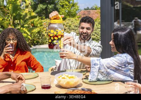 Verschiedene Freunde lachen und teilen sich ein erfrischendes rotes Getränk aus einem Krug mit Zitronen, genießen ein sonniges Mittagessen im Freien und einen entspannten Sommer Stockfoto