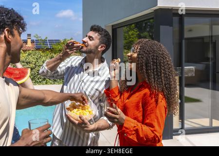 Gruppe verschiedener Freunde, die eine sonnige Poolparty im Hinterhof genießen, lachen und Pizza, Chips und Wassermelone teilen, während sie sich in einem warmen Sommer entspannen Stockfoto