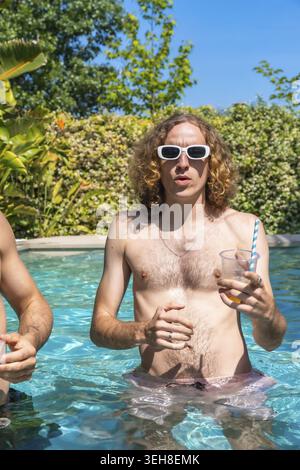Junger Mann mit lockigen Haaren und weißer Sonnenbrille, der an einem warmen Sommertag einen erfrischenden Drink in einem Swimmingpool genießt, Urlaub und Entspannung erlebt Stockfoto