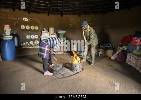 Zwei Menschen um ein brennendes Feuer in einer traditionellen Hütte, umgeben von Haushaltsgegenständen, Königreich Lesotho, Lesotho Stockfoto
