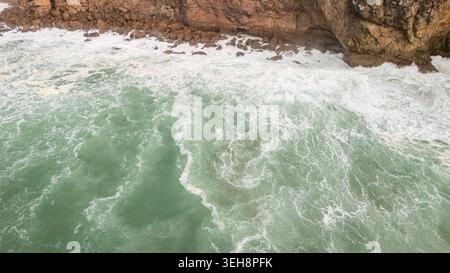 Aus der Vogelperspektive der rauen türkisfarbenen Meereswellen, die gegen die zerklüfteten Kalksteinklippen mit weißem Schaum in Sagres, Faro, Portugal, krachen. Stockfoto
