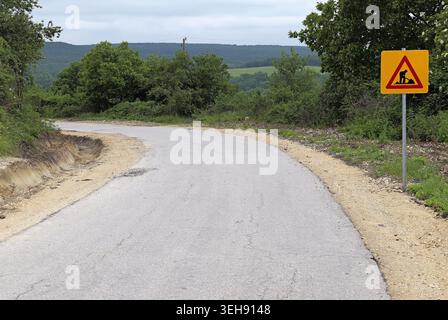Gelbes Schild für die Straßenbauarbeiten, auf dem ein Arbeiter grabt und Fahrer vor laufenden Bauarbeiten oder Wartungsarbeiten auf einer kurvenreichen Asphaltstraße gewarnt werden Stockfoto