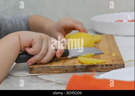 Kinderhände schneiden gekochte Kartoffeln auf Holzbrett mit Messer, Kartoffeln in kleine Stücke geschnitten, Hausküche Aktivität, Kochkunst lernen, einfach Stockfoto