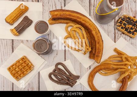 Verschiedene spanische gebratene Gebäckstücke wie Churros, dicke porras und Waffeln werden mit heißer Schokolade auf einer weißen Holzoberfläche serviert. Stockfoto