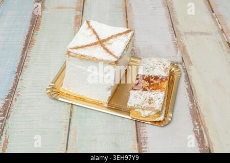 Ein quadratischer Milhojas-Kuchen mit weißer Glasur und Zimtkreuz sitzt auf einem goldenen Tablett über einem grünen Holztisch. Stockfoto