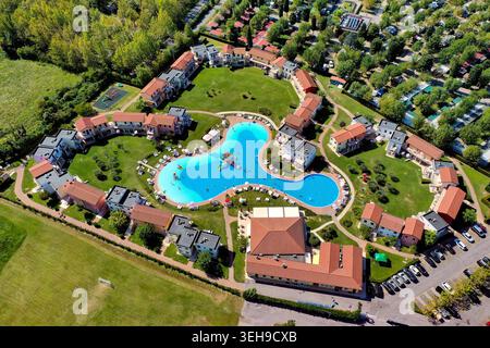 Aus der Vogelperspektive auf einen Resortkomplex mit Menschen, die in einem großen blauen Pool schwimmen, umgeben von bunten Häusern und grünen Rasenflächen in der Nähe des Gardasees. Stockfoto