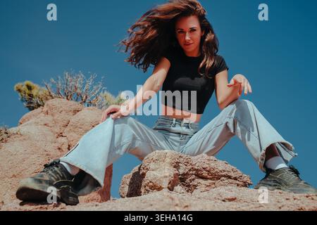 Frau mit lockigen Haaren, die auf einem Felsen in der Steppe sitzt, mit flachem Blick, Turnschuhen und Jeans Stockfoto